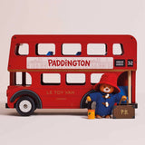A toy Paddington Bear in a red hat and blue coat stands beside a wooden red London double-decker bus.