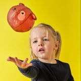 A young girl with blonde hair in pigtails, wearing a black sweater, tosses a red ball with a face design. The background is bright yellow, creating a playful and cheerful atmosphere.