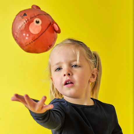 A young girl with blonde hair in pigtails, wearing a black sweater, tosses a red ball with a face design. The background is bright yellow, creating a playful and cheerful atmosphere.