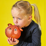 A young girl in a black shirt gently blows into a bear-shaped balloon on a bright yellow background. Her expression is focused and playful.