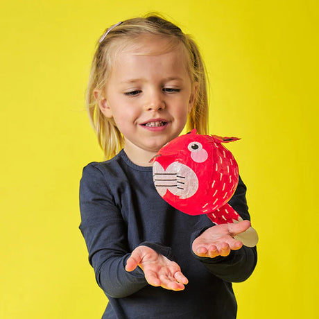 A young child with blonde hair smiles while holding a red, fish-shaped craft. The vibrant yellow background adds a cheerful, playful tone.