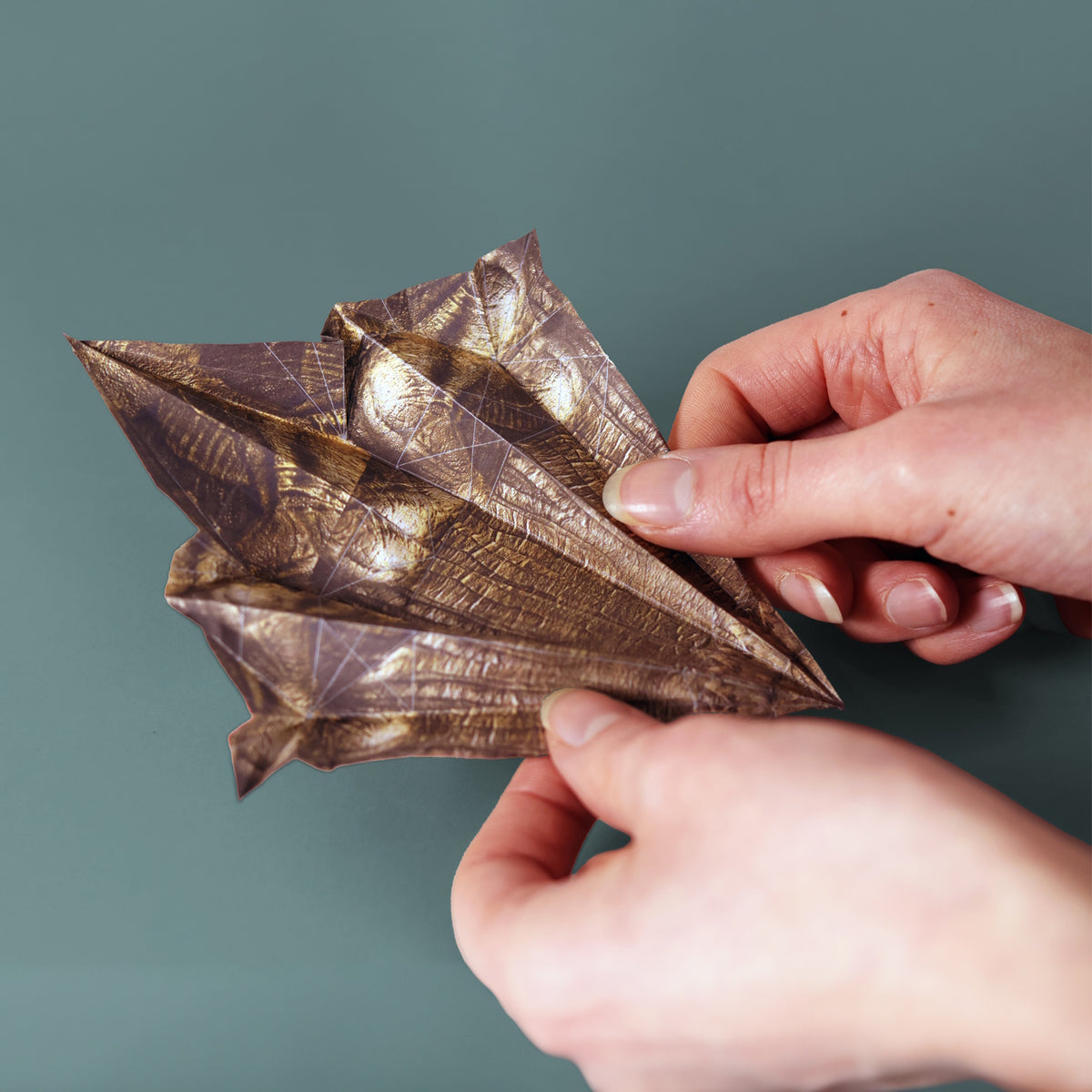 Hands folding a metallic brown patterned paper dinosaur model against a plain background.