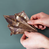 Hands folding a metallic brown patterned paper dinosaur model against a plain background.