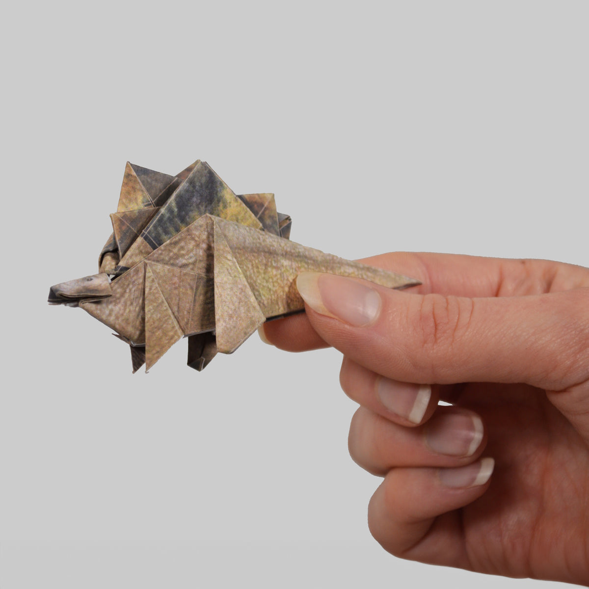 A hand holding a folded origami stegosaurus dinosaur made from pattern-printed paper.