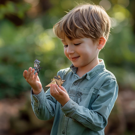 Young boy holding and looking at two small origami dinosaurs outdoors.