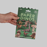 Hand holding the green Paper Dinos origami kit package against a neutral grey background.