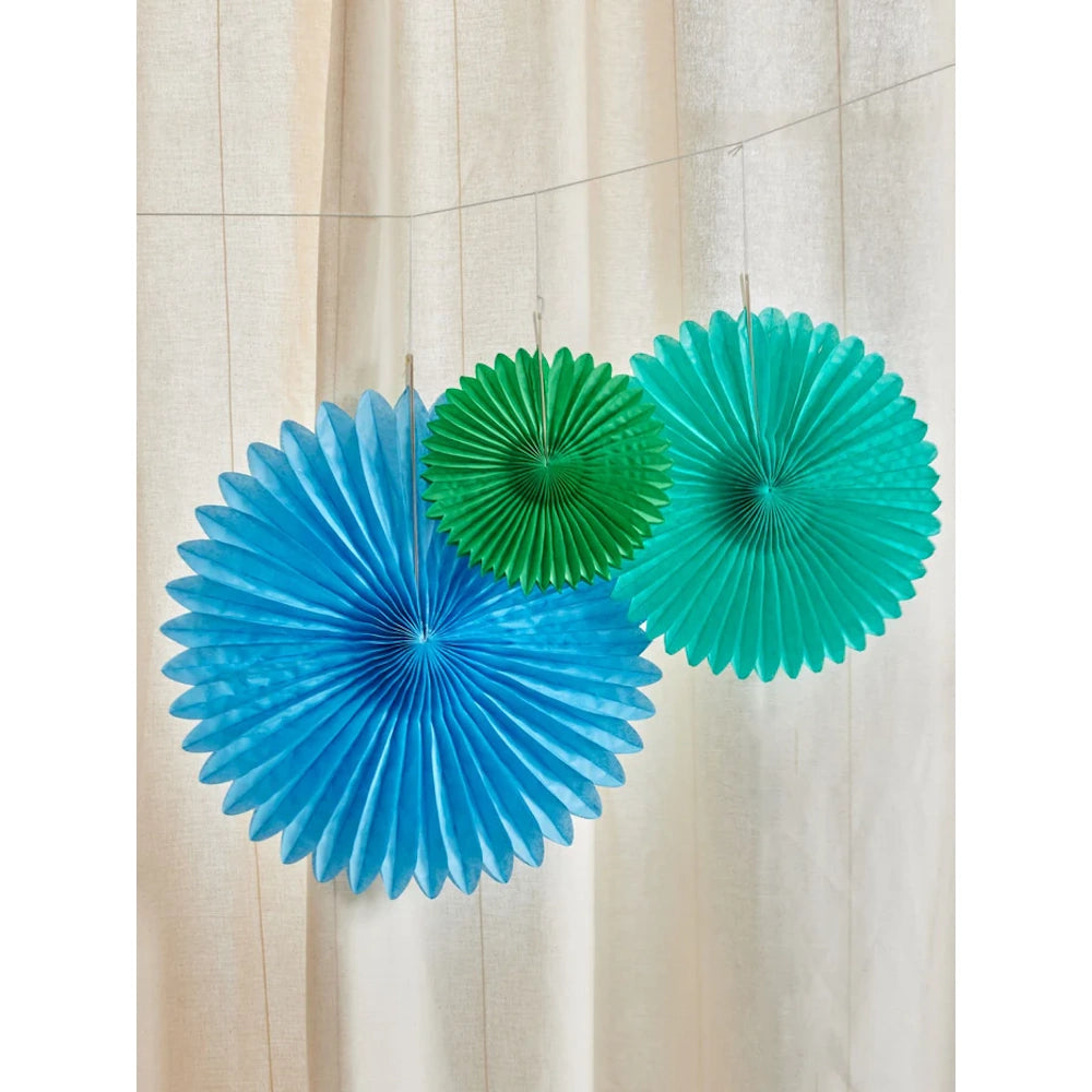 Three colorful paper fans hang against a light curtain. From left to right are blue, green, and teal fans, creating a festive and cheerful atmosphere.