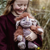 A smiling young girl holding a plush grey monster toy with a brown mane, single horn, and large floppy ears.
