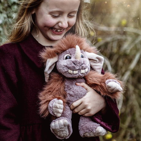 A smiling young girl holding a plush grey monster toy with a brown mane, single horn, and large floppy ears.