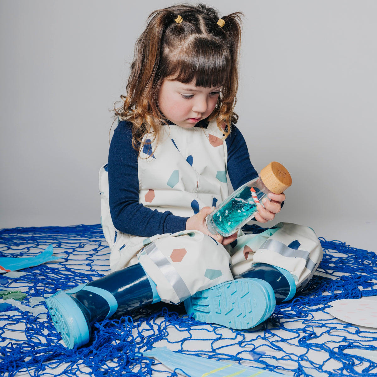 A young girl in patterned overalls and blue boots sits on a blue net, holding a sensory bottle.