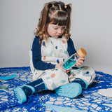 A young girl in patterned overalls and blue boots sits on a blue net, holding a sensory bottle.