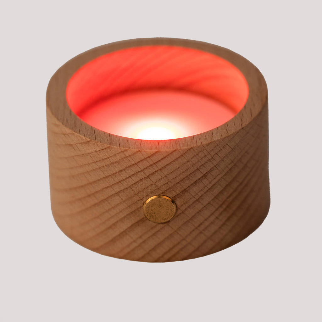 A round wooden candle holder with a glowing red LED light inside. The wood surface has a natural grain and a brass button accent. The mood is warm and soothing.
