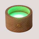A round wooden candle holder with a glowing red LED light inside. The wood surface has a natural grain and a brass button accent. The mood is warm and soothing.