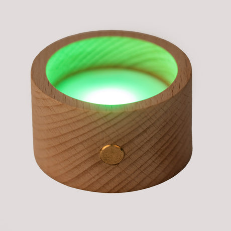 A round wooden candle holder with a glowing red LED light inside. The wood surface has a natural grain and a brass button accent. The mood is warm and soothing.