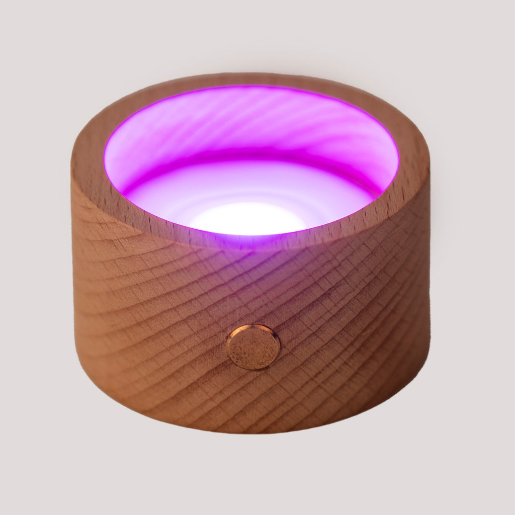 A round wooden candle holder with a glowing red LED light inside. The wood surface has a natural grain and a brass button accent. The mood is warm and soothing.
