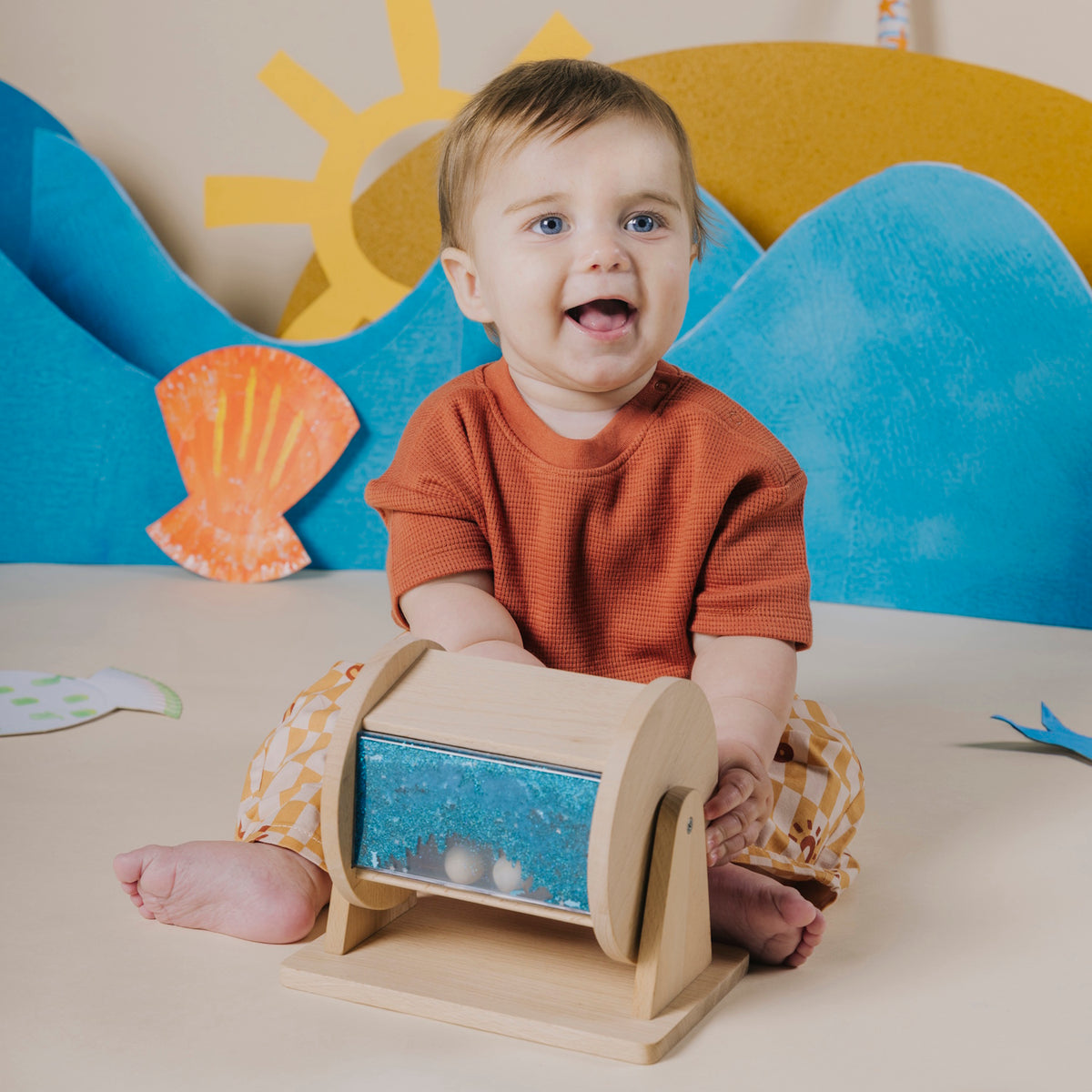 Baby sitting up and smiling while playing with a wooden sensory roller.