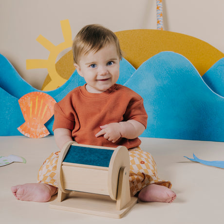 Baby sitting on the floor and pointing at a wooden sensory roller.