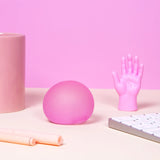 Pink round squishy stress ball on a desk alongside a small plastic hand figurine and office items.