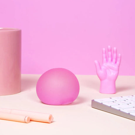 Pink round squishy stress ball on a desk alongside a small plastic hand figurine and office items.