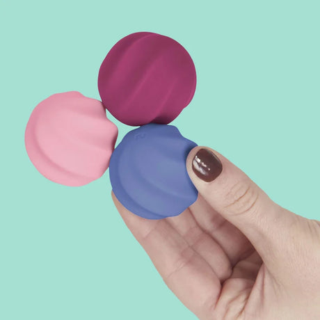 Hand holding three swirled Speks Odds fidget magnets in blue, pink, and magenta against a teal background.