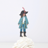 Pirate captain topper in a blue coat and feathered tricorn hat on a wooden pick in swirled white frosting.