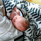 Sleeping newborn wrapped in a white muslin blanket with a black fern leaf pattern.