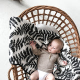 Baby relaxing in a wicker chair on a black-and-white fern printed muslin blanket.