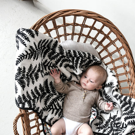 Baby relaxing in a wicker chair on a black-and-white fern printed muslin blanket.
