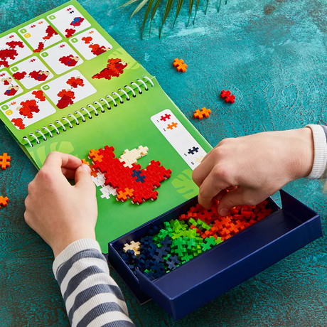 Child building a red dinosaur with Plus-Plus pieces on a green activity pad.