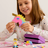 Young girl smiling while holding a colorful unicorn head made of Plus-Plus blocks, with a fairytale activity pad and toys.