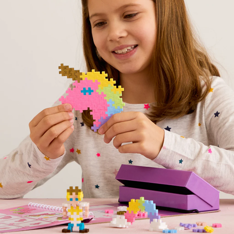 Young girl smiling while holding a colorful unicorn head made of Plus-Plus blocks, with a fairytale activity pad and toys.