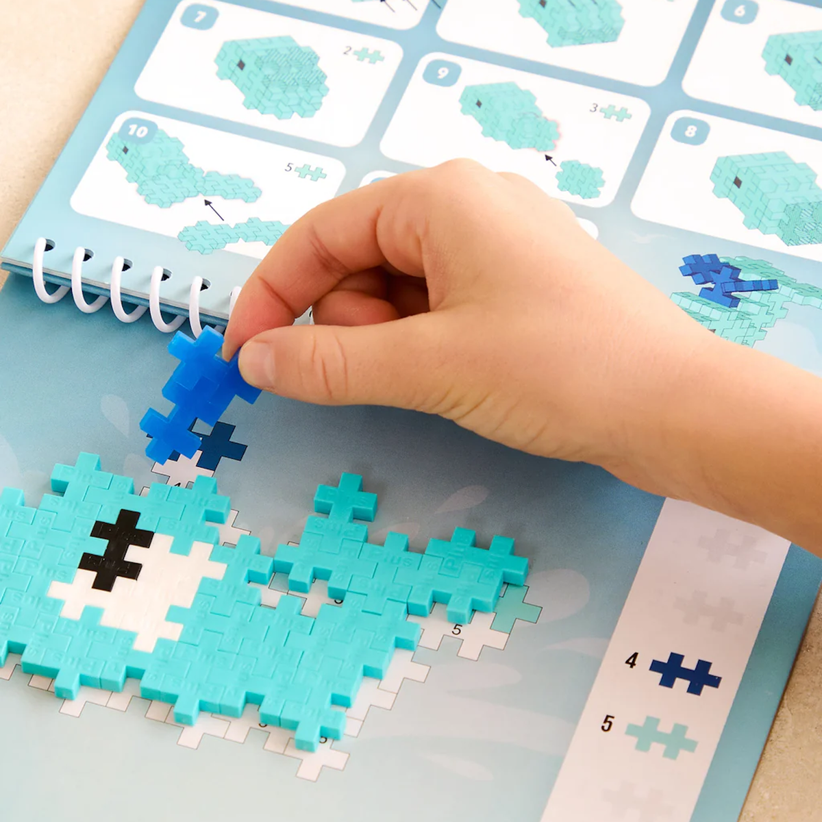 Child's hand placing a blue Plus-Plus piece on a whale puzzle in an ocean-themed activity book.