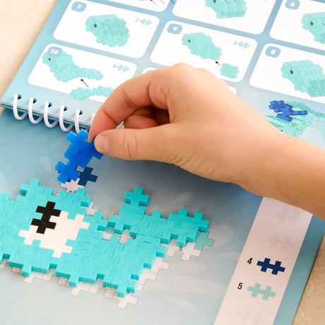 Child's hand placing a blue Plus-Plus piece on a whale puzzle in an ocean-themed activity book.