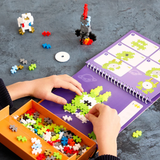 Child building a lime green alien on a purple Plus-Plus activity pad with a rocket and astronaut in the background.