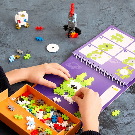 Child building a lime green alien on a purple Plus-Plus activity pad with a rocket and astronaut in the background.