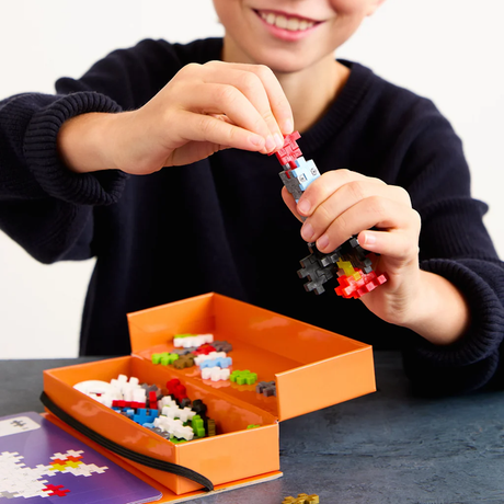 Child building a rocket with Plus-Plus blocks over an orange activity case and puzzle instructions.