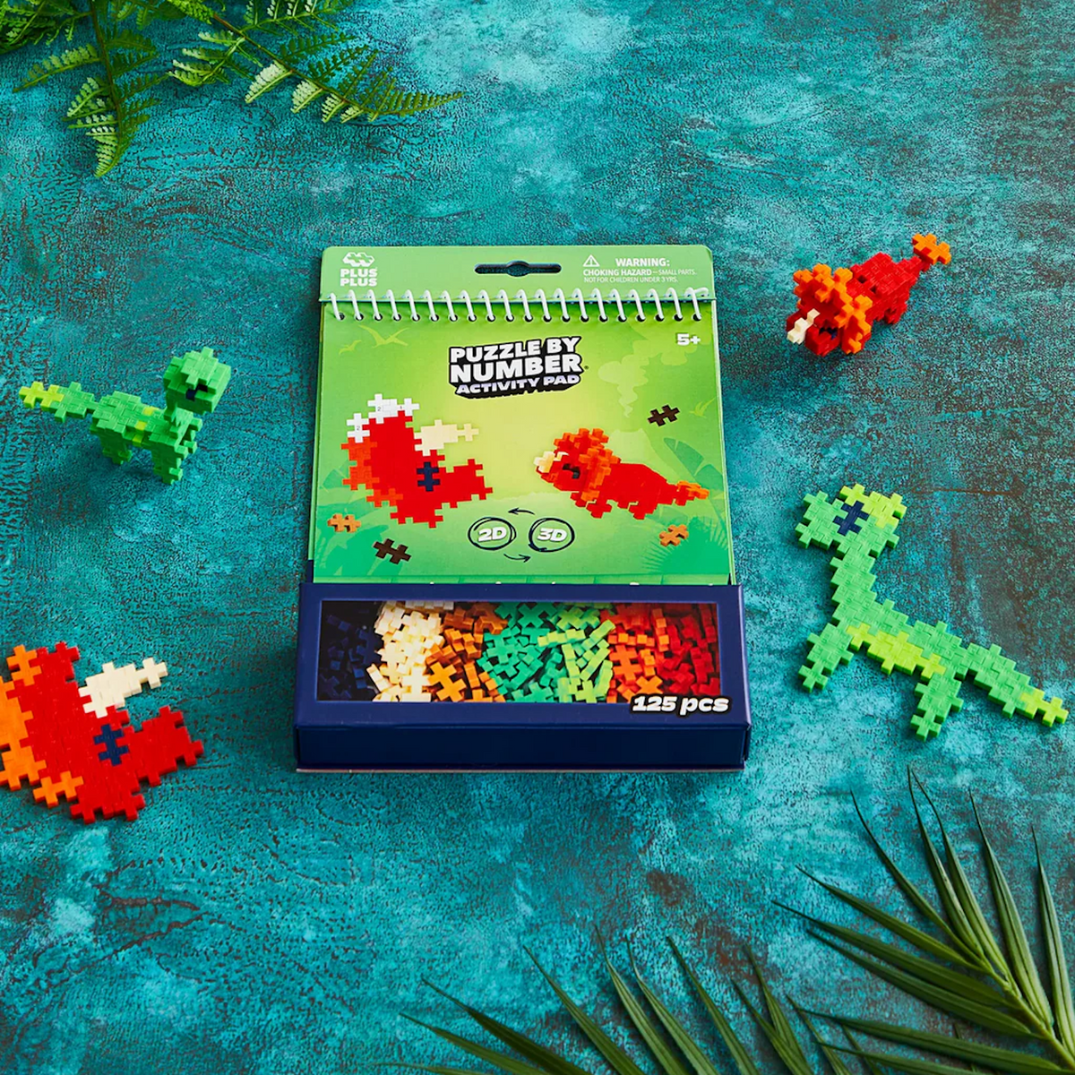 Plus-Plus Puzzle by Number Activity Pad with colorful 2D and 3D dinosaur builds and a 125-piece storage box.