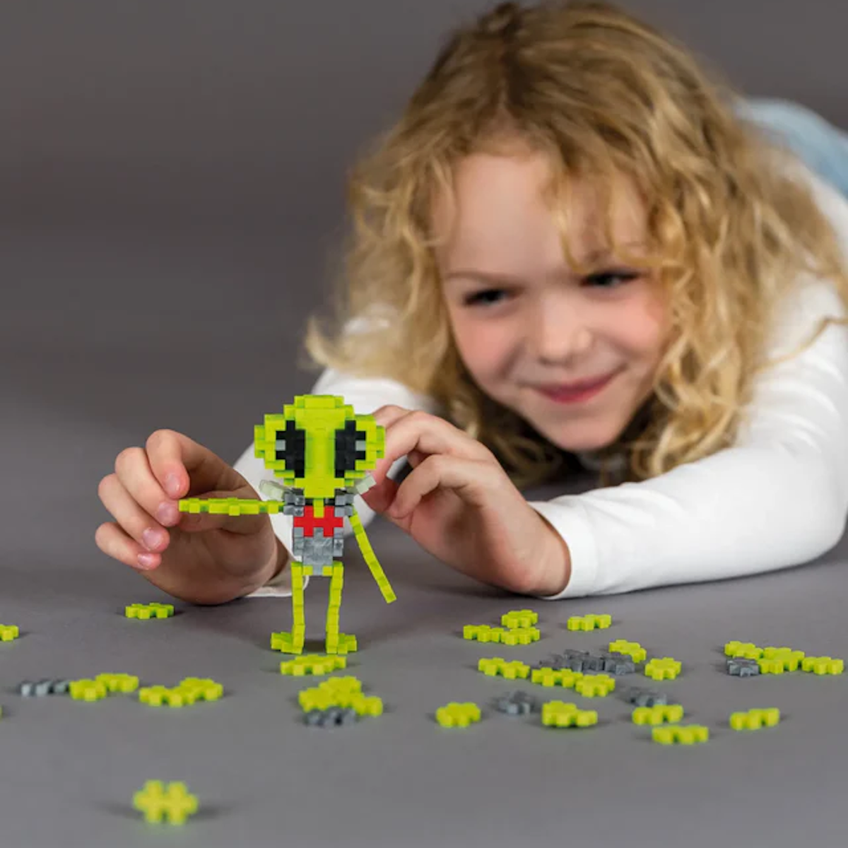 A young child with curly hair smiles while assembling a green alien figure from small building blocks on a gray surface, conveying creativity and joy.