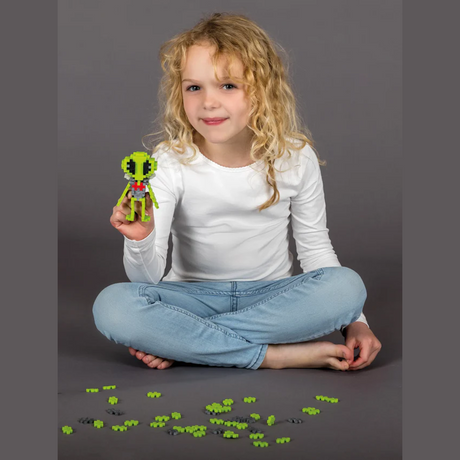 Young girl with curly hair sits cross-legged, holding a small green toy figure with black eyes, surrounded by scattered toy pieces, smiling softly.
