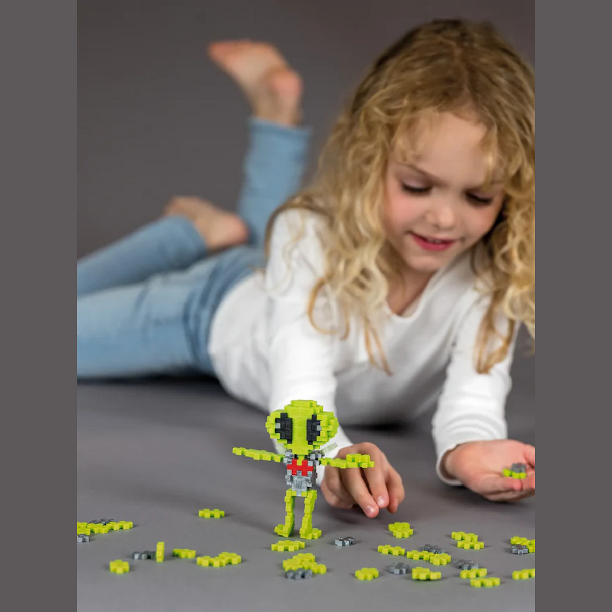 A young child with curly blonde hair, wearing a white shirt and jeans, lies on the floor, smiling while building a small green alien figure with toy blocks.