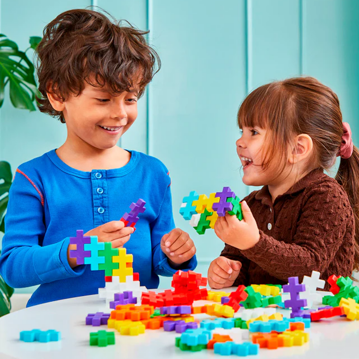 Two children smiling and playing with colorful interlocking building blocks at a table. The background features a light blue wall and a leafy plant.