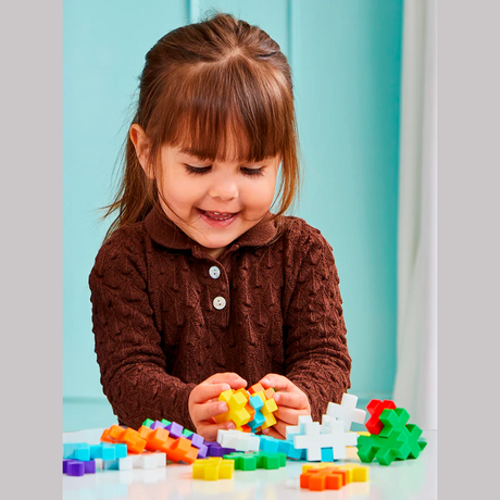 A young child with brown hair plays happily with colorful interlocking blocks at a white table. The scene feels joyful and focused, set against a teal background.