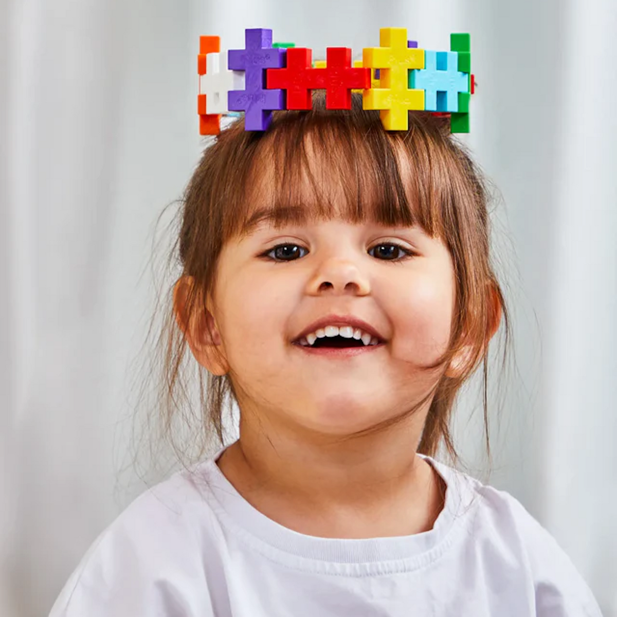 A joyful child wearing a colorful toy crown made of interlocking pieces smiles widely. The background is softly blurred, emphasizing her happy expression.