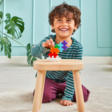 Smiling child with curly hair, wearing a striped shirt, builds with colorful plus-shaped blocks on a wooden stool. A large plant decorates the background.
