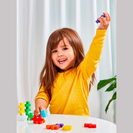 A joyful child with brown hair in a yellow sweater holds up a purple building block. She's surrounded by colorful blocks on a white table, smiling brightly.