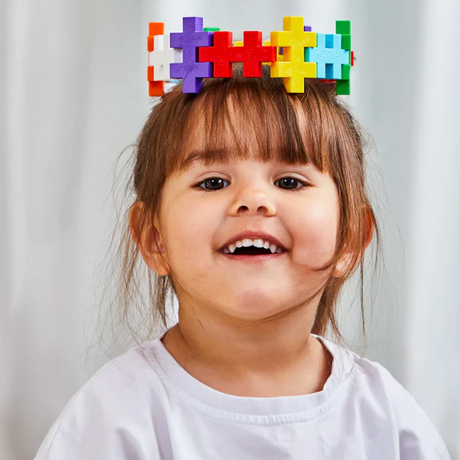 A smiling child wearing a crown made of colorful interlocking blocks; their white shirt contrasts the bright blocks, creating a playful and joyful mood.