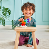 A joyful child with curly hair plays with colorful puzzle blocks on a wooden stool. The setting is a bright, cheerful room with a large plant.