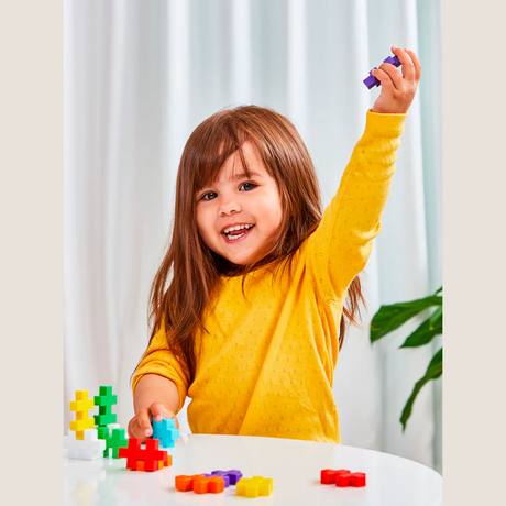 A smiling young girl in a yellow shirt raises a purple block triumphantly. She is playing with colorful toy blocks on a white table, exuding joy and excitement.