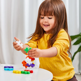 A young child with long brown hair, wearing a yellow sweater, smiles and builds a colorful block tower at a white table, conveying joy and focus.
