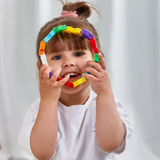 A smiling child with bangs and a ponytail holds a colorful ring of connected plastic blocks, looking playful and joyful against a soft background.